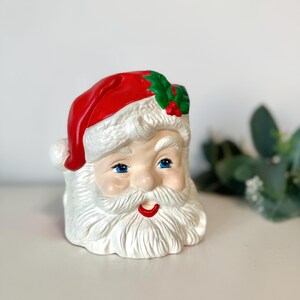 May include: A ceramic Santa Claus head with a red hat, white beard, and blue eyes. The hat is adorned with holly and berries. The Santa Claus head is a festive decoration.