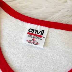 May include: A white t-shirt with a red collar trim featuring an Anvil brand tag. The tag reads: anvil, PRE-SHRUNK, 100% COTTON/COTON, -S-, MADE IN U.S.A., STYLED FOR WOMEN, OVER FOR CARE. The fabric is white with a visible weave and red stitching.