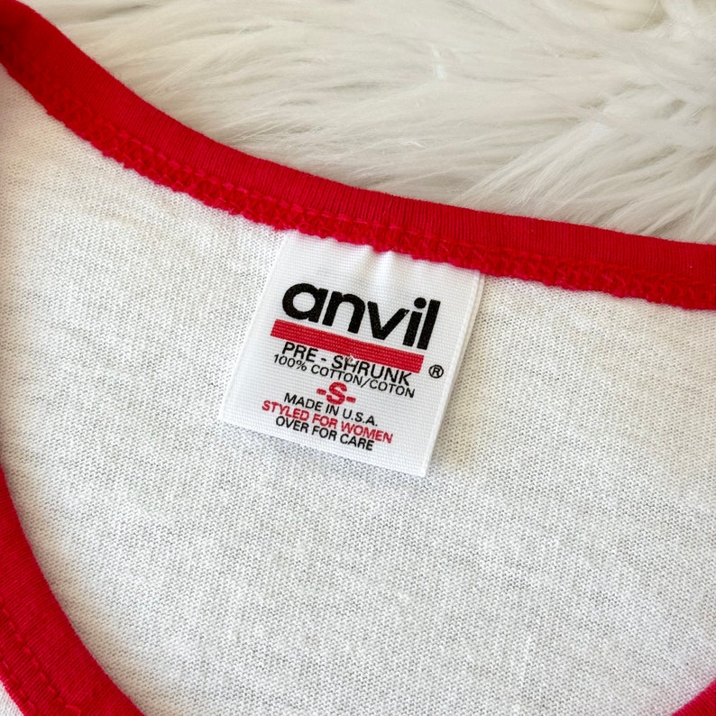 May include: A white t-shirt with a red collar trim featuring an Anvil brand tag. The tag reads: anvil, PRE-SHRUNK, 100% COTTON/COTON, -S-, MADE IN U.S.A., STYLED FOR WOMEN, OVER FOR CARE. The fabric is white with a visible weave and red stitching.