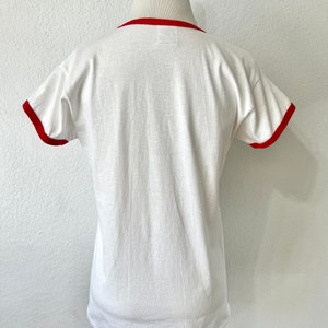 May include: A white short-sleeve ringer t-shirt featuring a solid red crew neckline and matching red trim on the sleeve cuffs. The garment is displayed on a mannequin, showing a plain back view with a smooth fabric texture and a standard hemline.