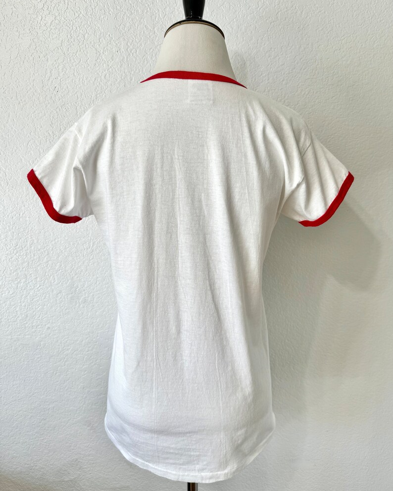 May include: A white short-sleeve ringer t-shirt featuring a solid red crew neckline and matching red trim on the sleeve cuffs. The garment is displayed on a mannequin, showing a plain back view with a smooth fabric texture and a standard hemline.