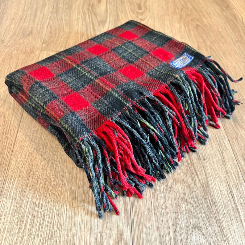 Puede incluir: Una manta a cuadros roja, negra y verde doblada con flecos rojos. La manta tiene un patr&oacute;n rectangular con una peque&ntilde;a etiqueta azul en la esquina. La manta est&aacute; hecha de un material suave.