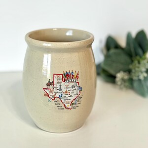 May include: A beige ceramic crock with a speckled finish featuring a colorful map of Texas. The graphic includes the text Texas and The Lone Star State, along with various illustrations of landmarks, animals, and flags across the state map.