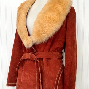 May include: A rust-colored suede coat with a tan faux fur collar and a matching belt. The coat features tan stitching details and is displayed on a mannequin. A vintage-inspired outerwear piece.