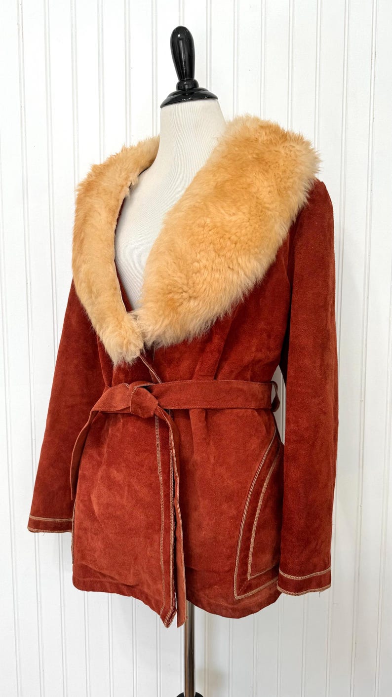 May include: A rust-colored suede coat with a tan faux fur collar and a matching belt. The coat features tan stitching details and is displayed on a mannequin. A vintage-inspired outerwear piece.