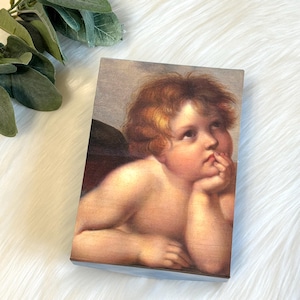May include: A rectangular wooden box featuring a Renaissance-style painting of a cherubic figure with curly red hair, resting its chin on its hand. The artwork has a soft, warm color palette.