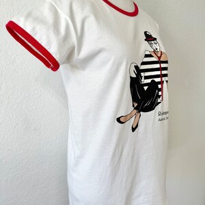 May include: A white ringer t-shirt with red trim on the collar and sleeves. The front features a graphic of a person in a black and white striped shirt, black pants, and a bowler hat. The text below the graphic reads Shakespeare and Ashland, Oregon.