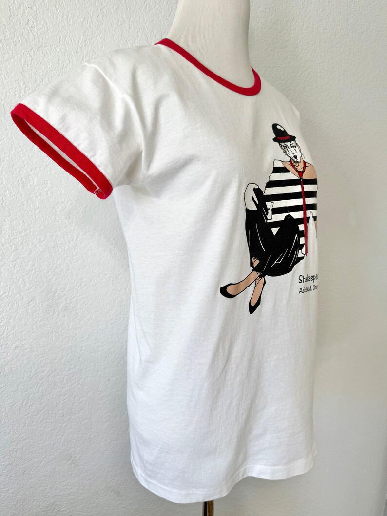 May include: A white ringer t-shirt with red trim on the collar and sleeves. The front features a graphic of a person in a black and white striped shirt, black pants, and a bowler hat. The text below the graphic reads Shakespeare and Ashland, Oregon.