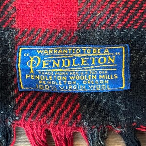 Puede incluir: Primer plano de una bufanda a cuadros roja, negra y azul con flecos. Una etiqueta azul con texto dorado dice "WARRANTED TO BE A PENDLETON" y "100% VIRGIN WOOL". La bufanda es de lana.