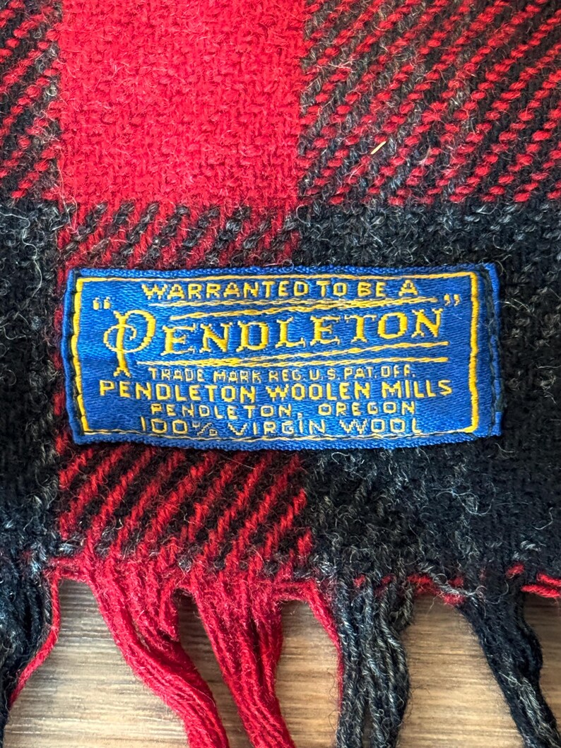 Puede incluir: Primer plano de una bufanda a cuadros roja, negra y azul con flecos. Una etiqueta azul con texto dorado dice "WARRANTED TO BE A PENDLETON" y "100% VIRGIN WOOL". La bufanda es de lana.