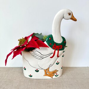 May include: A white ceramic goose planter with a green wreath and red bow around its neck. The planter is decorated with red and green accents and holds red poinsettia flowers. The goose has an orange beak and feet.