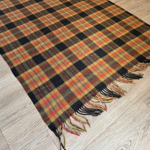 May include: A rectangular plaid blanket in shades of black, orange, yellow, and red. The blanket has a fringed edge and is displayed on a light wood surface. The plaid pattern is a classic design, suitable for home decor.