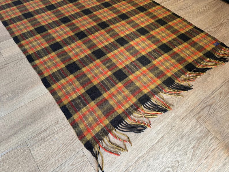 May include: A rectangular plaid blanket in shades of black, orange, yellow, and red. The blanket has a fringed edge and is displayed on a light wood surface. The plaid pattern is a classic design, suitable for home decor.