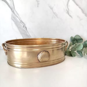 May include: An oval-shaped brass basin featuring a polished metallic finish and a decorative embossed seashell motif centered on the front exterior. The vessel includes two small side handles and a smooth, rounded rim, presenting a clean design.