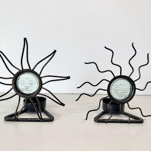 May include: Two black metal candle holders featuring a sun design with radiating wire rays. Each holder has a central circular glass insert embossed with a human face and a triangular base with a small cylindrical cup for holding a candle.