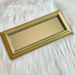 May include: A rectangular mirror featuring a wide, textured gold frame with a speckled finish. The frame has a beveled inner edge that transitions into a smooth, reflective glass surface. The overall design is elongated and horizontal in orientation.