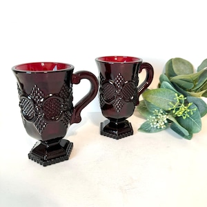 May include: Two ruby red vintage glass mugs with ornate floral and geometric patterns. Each mug has a handle and sits on a square base. The interior of the mugs is a vibrant red, and they are displayed on a white surface.