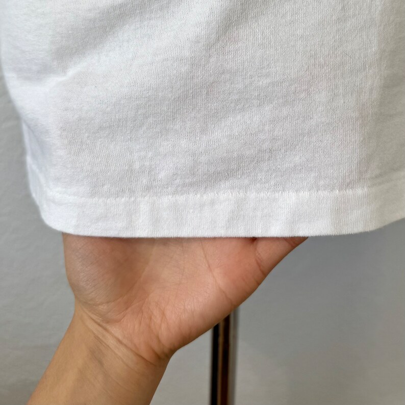 May include: A plain white t-shirt featuring a standard hemline with visible stitching along the bottom edge. The fabric has a smooth, opaque texture and a clean, minimalist appearance, showcasing the quality of the material and the construction.