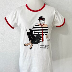 May include: A white ringer t-shirt with red trim on the collar and sleeves. The front features a graphic of a person in a black and white striped shirt, black pants, a red tie, and a black hat. The text reads Shakespeare Ashland, Oregon.