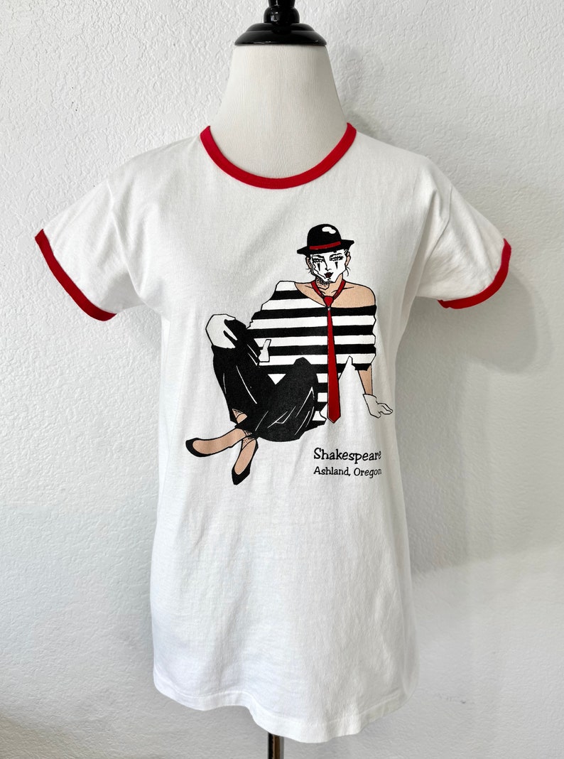 May include: A white ringer t-shirt with red trim on the collar and sleeves. The front features a graphic of a person in a black and white striped shirt, black pants, a red tie, and a black hat. The text reads Shakespeare Ashland, Oregon.