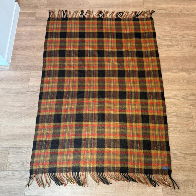 May include: A rectangular plaid blanket in shades of black, gold, and red. The blanket has a fringed edge and a classic tartan pattern. This cozy blanket is suitable for indoor or outdoor use.