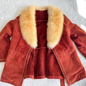 May include: A rust-colored suede coat with a cream-colored fur collar. The coat has a matching belt and a soft, plush interior. The coat is open, revealing the interior lining. The coat is laid out on a white surface.