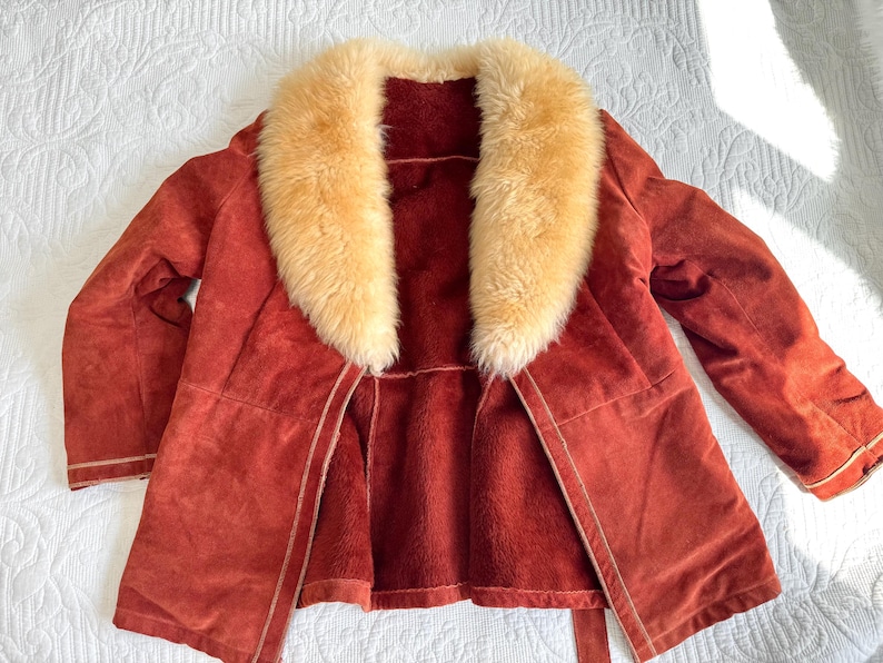 May include: A rust-colored suede coat with a cream-colored fur collar. The coat has a matching belt and a soft, plush interior. The coat is open, revealing the interior lining. The coat is laid out on a white surface.