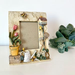 May include: A decorative picture frame with a garden theme. The frame features a textured beige border with a butterfly, birdhouse, tulips, and a watering can. The frame is approximately 8 inches tall and 6 inches wide.