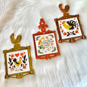 May include: Three vintage trivets with rooster and floral designs. The trivets have a square tile center with a decorative frame and a rooster at the top. One trivet has the text "The Cheese Table". The frames are gold, brown, and orange.