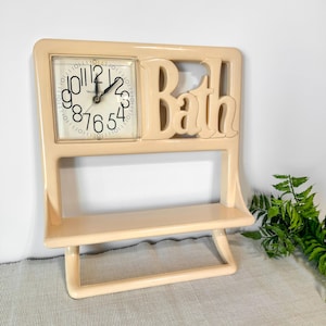 May include: A beige-colored clock and shelf unit with the word "Bath" cut out at the top. The clock has a white face with black numbers and hands. The shelf is rectangular and the unit has a stand.