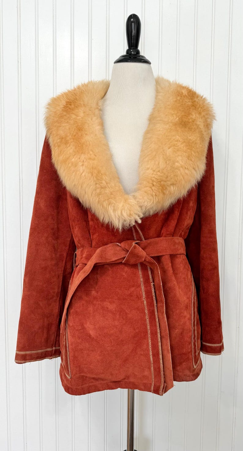 May include: Rust-colored suede coat with a tan faux fur collar and a matching belt. The coat has a wrap-style design with visible stitching and is displayed on a mannequin. A vintage outerwear piece.