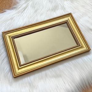 May include: A rectangular gold-framed mirror with a reflective surface. The frame has a detailed, ornate design with a beaded inner edge. The mirror is set against a white, fluffy background.
