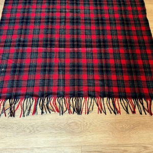 Puede incluir: Bufanda rectangular a cuadros rojos, negros y verdes con flecos en el borde inferior. La bufanda est&aacute; hecha de un material suave y est&aacute; colocada sobre una superficie de madera.