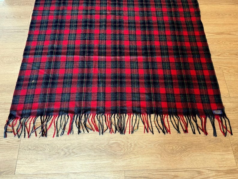 Puede incluir: Bufanda rectangular a cuadros rojos, negros y verdes con flecos en el borde inferior. La bufanda est&aacute; hecha de un material suave y est&aacute; colocada sobre una superficie de madera.