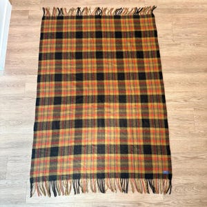 May include: A rectangular plaid blanket with a fringed edge. The blanket features a repeating pattern of black, orange, and gold squares. The blanket is made of a soft material and is suitable for use as a throw or a picnic blanket.