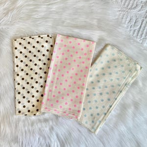 May include: Three folded cloths with polka dot patterns. One cloth has brown and gray dots, another has pink dots, and the third has blue dots. All are on a white, fluffy surface.