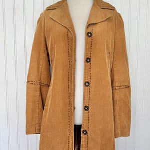 May include: A tan corduroy coat with a notched collar and long sleeves. The coat features black stitching details along the seams and around the decorative buttons. The coat is buttoned up and displayed on a mannequin.