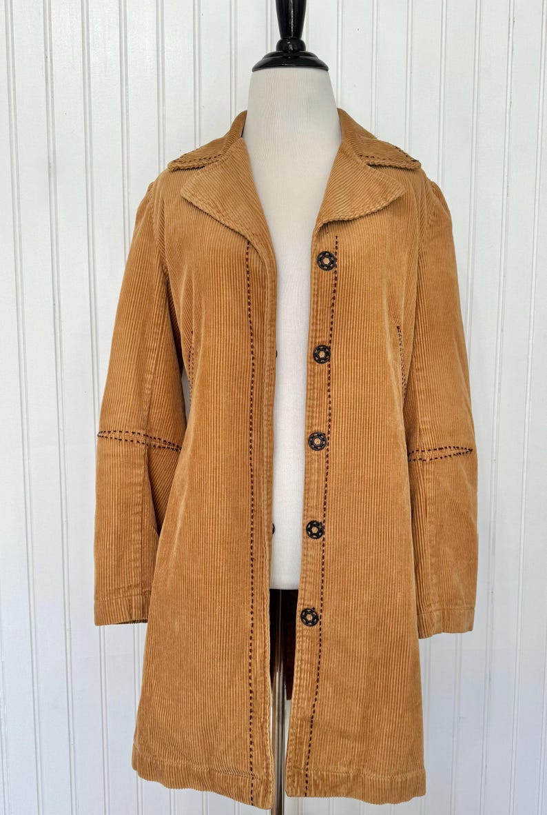 May include: A tan corduroy coat with a notched collar and long sleeves. The coat features black stitching details along the seams and around the decorative buttons. The coat is buttoned up and displayed on a mannequin.