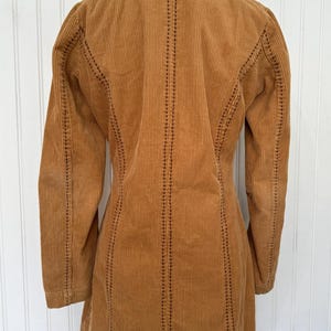 May include: A tan corduroy jacket with a notched collar and long sleeves. The jacket features vertical corduroy ribs and decorative stitching along the seams. The jacket is displayed on a black mannequin.
