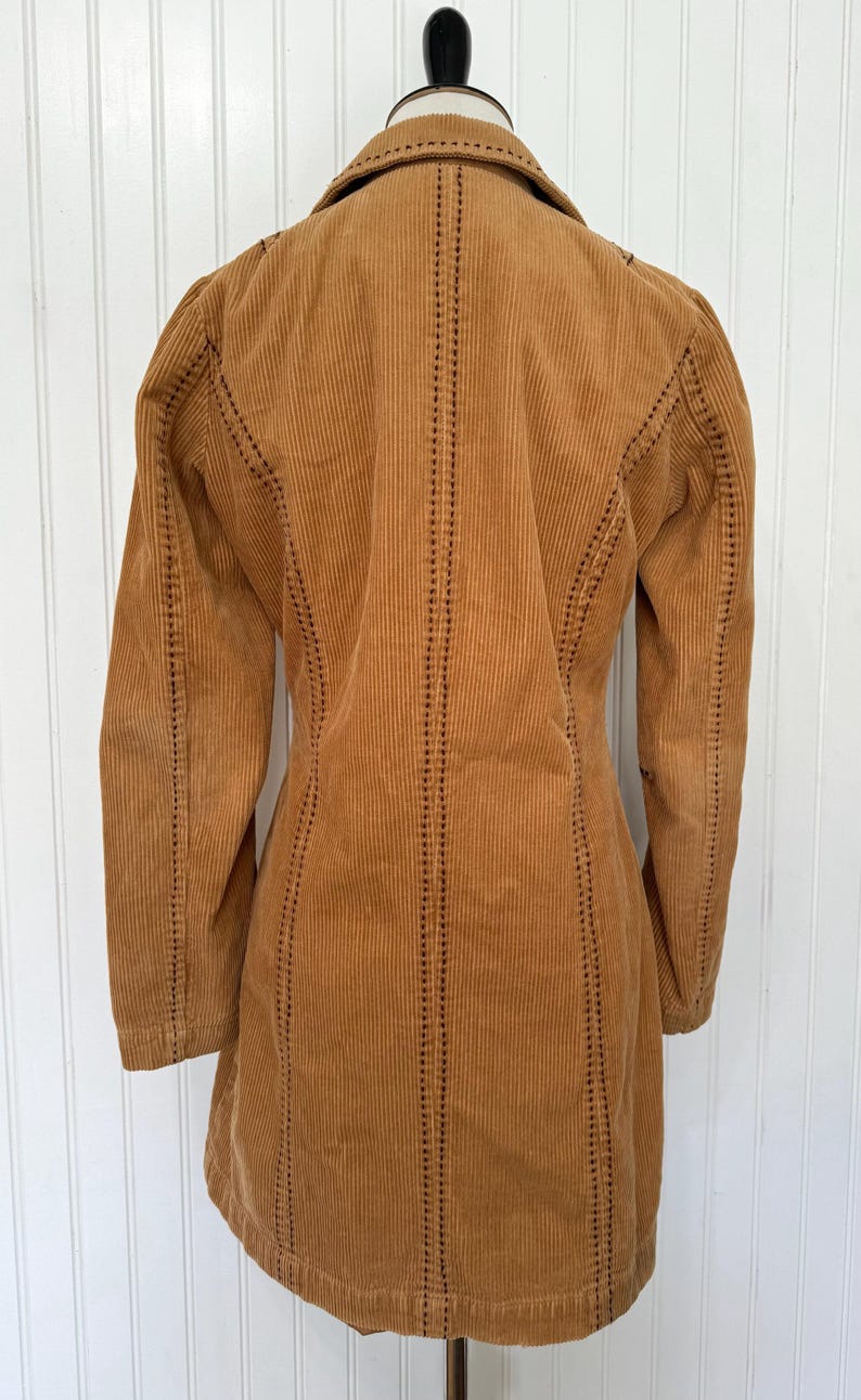 May include: A tan corduroy jacket with a notched collar and long sleeves. The jacket features vertical corduroy ribs and decorative stitching along the seams. The jacket is displayed on a black mannequin.
