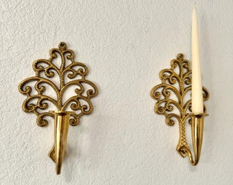 May include: Two ornate, gold-colored wall sconces with a scrolling leaf design. One sconce holds a tall, white candle. The sconces are mounted on a textured, off-white wall. Decorative home decor.