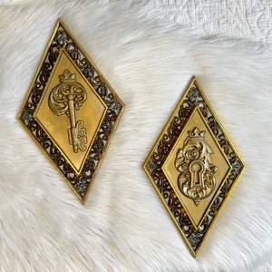 May include: Two gold-colored diamond-shaped wall decor pieces with ornate frames. One features a key with a crown and the other features a keyhole with a crown.