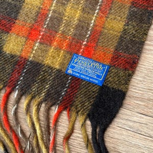 May include: A Pendleton scarf with a plaid pattern in shades of brown, orange, yellow, and black. The scarf has a fringed edge and a blue label that reads "Pendleton Woolen Mills, 100% Virgin Wool."