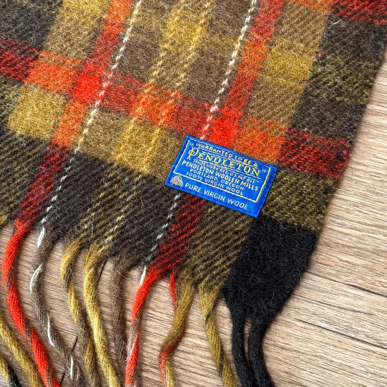 May include: A Pendleton scarf with a plaid pattern in shades of brown, orange, yellow, and black. The scarf has a fringed edge and a blue label that reads "Pendleton Woolen Mills, 100% Virgin Wool."
