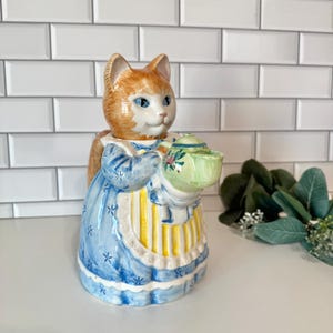 May include: A ceramic teapot shaped like an orange and white cat wearing a blue dress with a yellow striped apron. The cat holds a small, light green teapot with a floral design in its paws. The piece features a glossy finish and detailed painting.