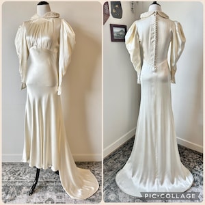 May include: Ivory-colored vintage wedding dress with a long train. The dress features a high collar with lace trim, long sleeves with puffed shoulders, and a fitted bodice. The back of the dress has a row of buttons. The dress is displayed on a mannequin.