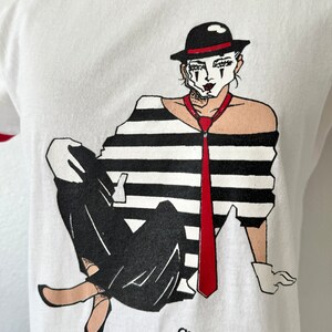 May include: A white ringer t-shirt with a red collar and sleeve trim. The graphic features a mime wearing a black hat, a black and white striped shirt, a red tie, and white gloves. The text below the illustration reads Shakespeare Ashland, Oregon.