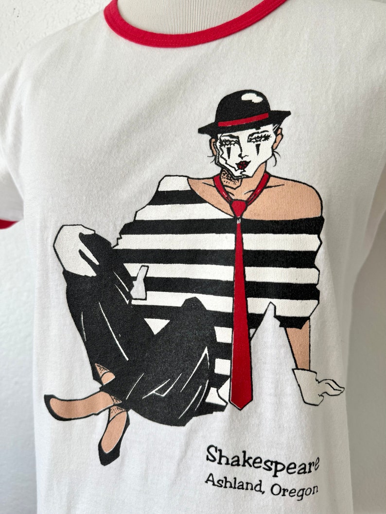 May include: A white ringer t-shirt with a red collar and sleeve trim. The graphic features a mime wearing a black hat, a black and white striped shirt, a red tie, and white gloves. The text below the illustration reads Shakespeare Ashland, Oregon.