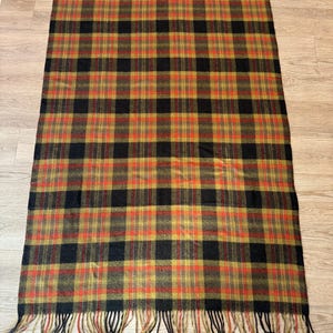 May include: A rectangular plaid blanket with a repeating pattern of black, orange, and gold squares. The blanket has a fringe of black and gold threads along the top and bottom edges. The blanket is lying on a wooden surface.