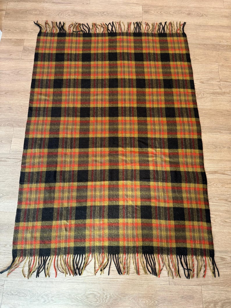 May include: A rectangular plaid blanket with a repeating pattern of black, orange, and gold squares. The blanket has a fringe of black and gold threads along the top and bottom edges. The blanket is lying on a wooden surface.